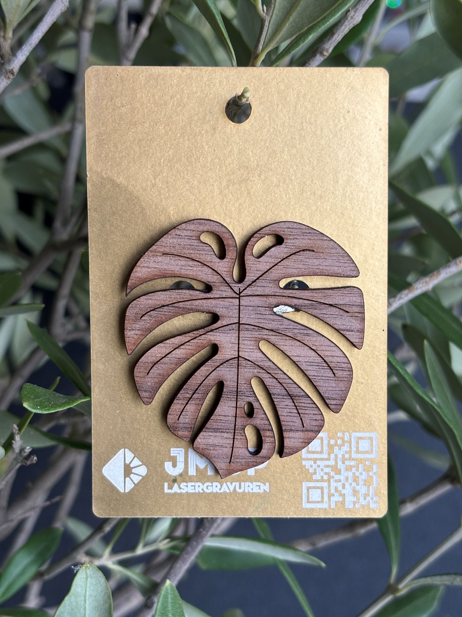 Holz-Lesezeichen in Form eines Monstera-Blattes mit Lasergravur auf goldenem Hintergrund vor grünen Blättern.